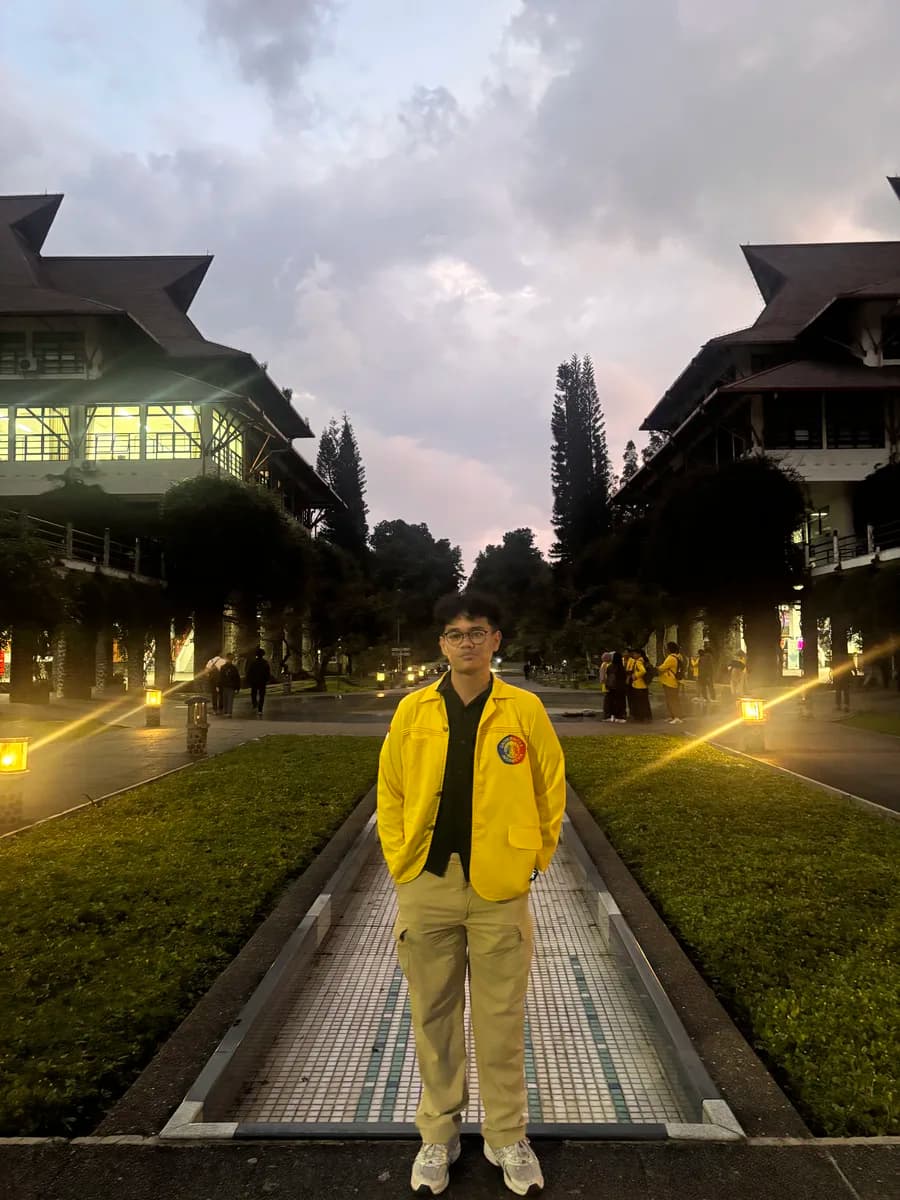 Wahyu wearing a yellow university jacket on campus at dusk